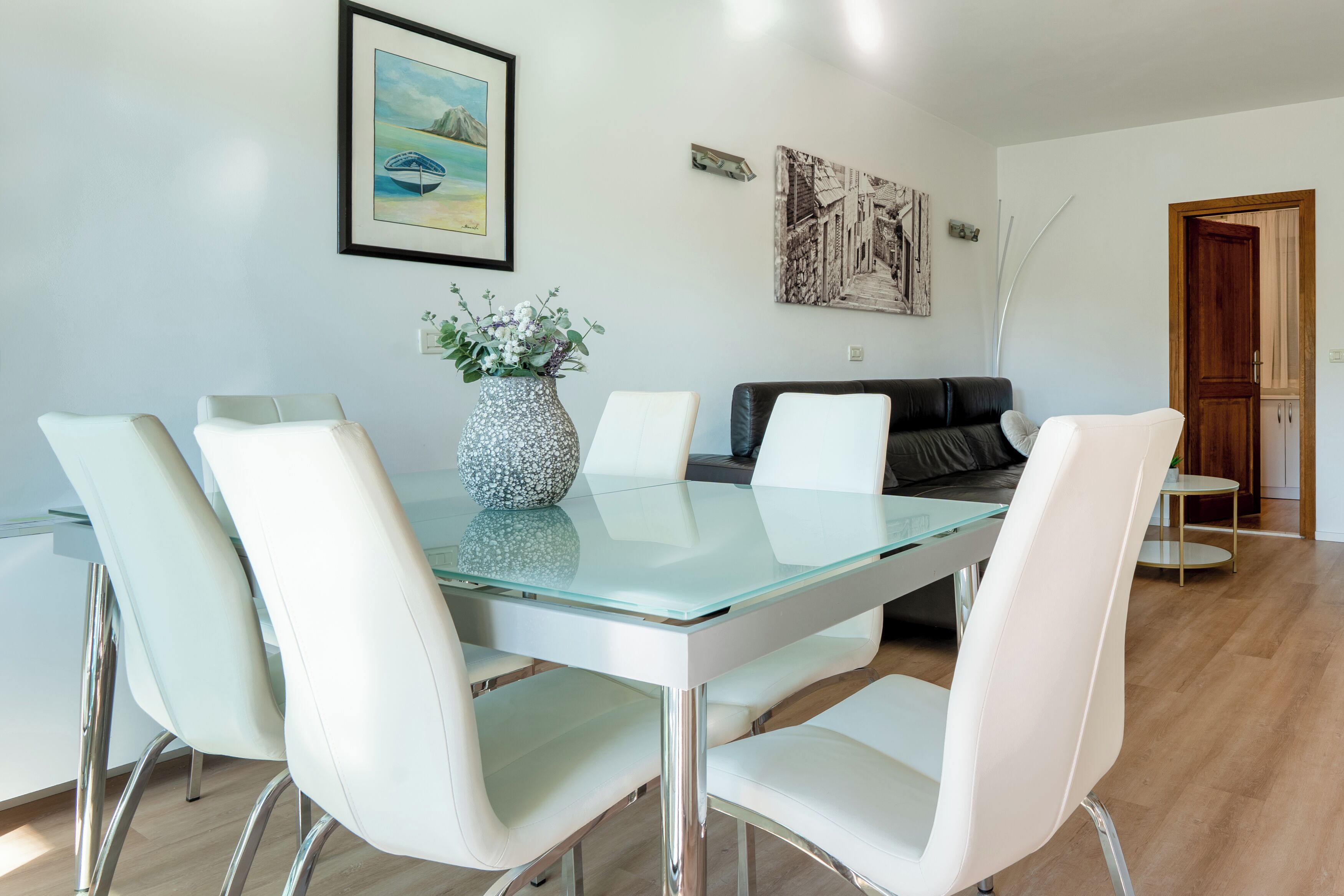 A modern dining area with a glass table and white chairs, a vase with flowers, and artwork on the walls.