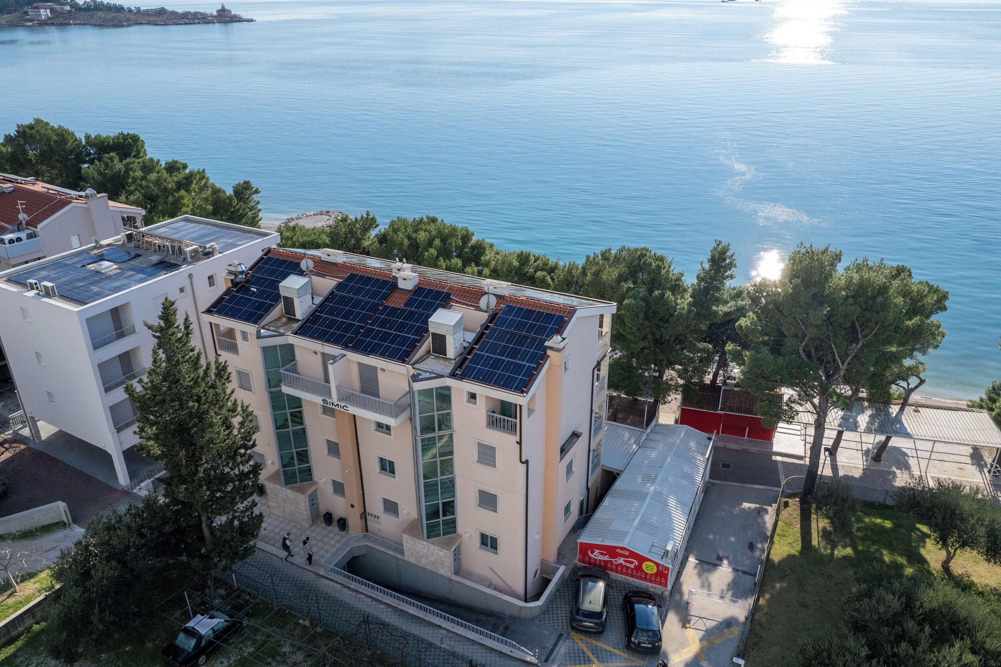A modern building with solar panels on the roof, a parking area, and a view of the sea.