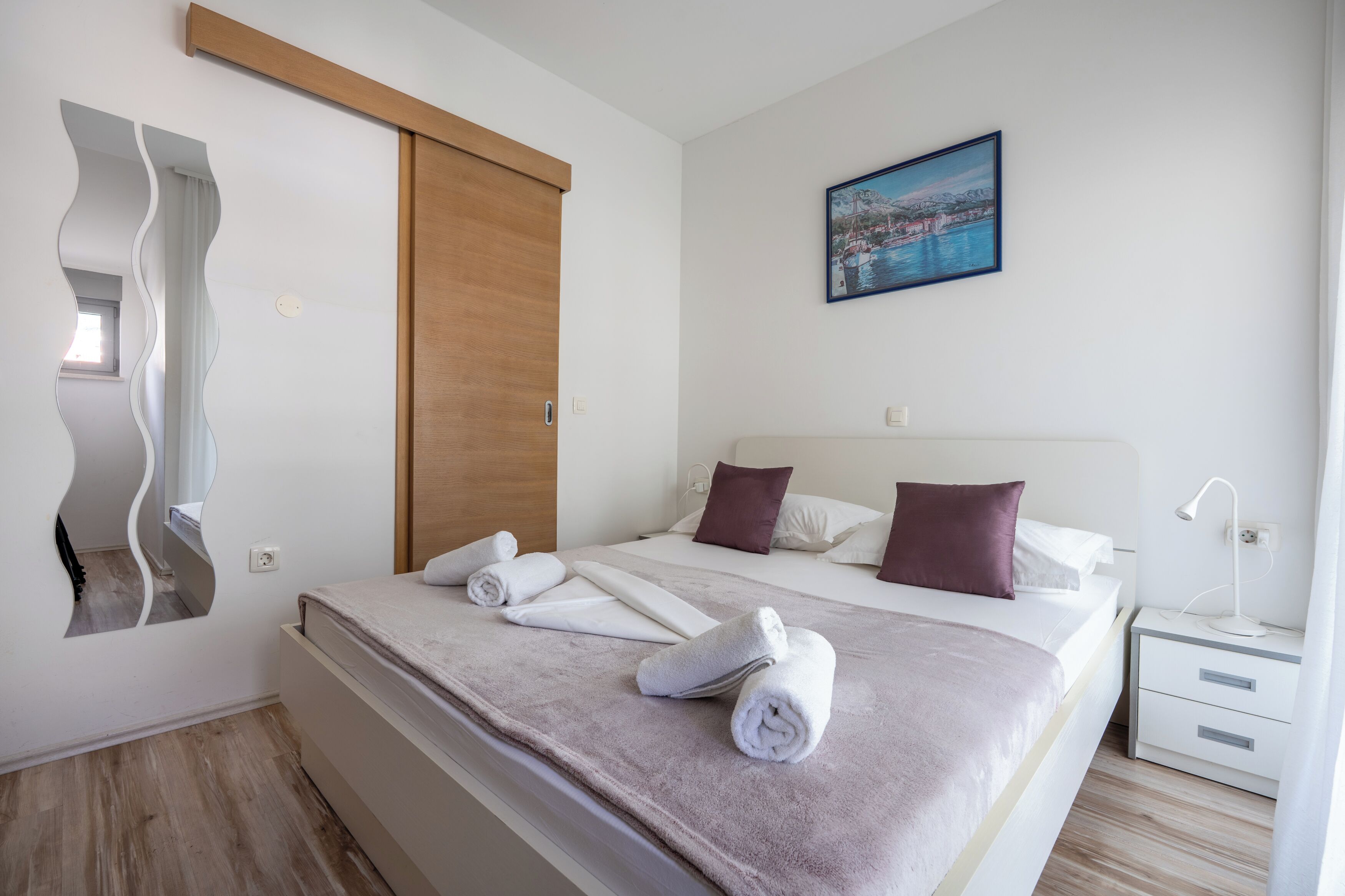 Camera d'albergo moderna con un letto grande, una porta scorrevole in legno, un quadro raffigurante una costa e uno specchio con motivo ondulato.