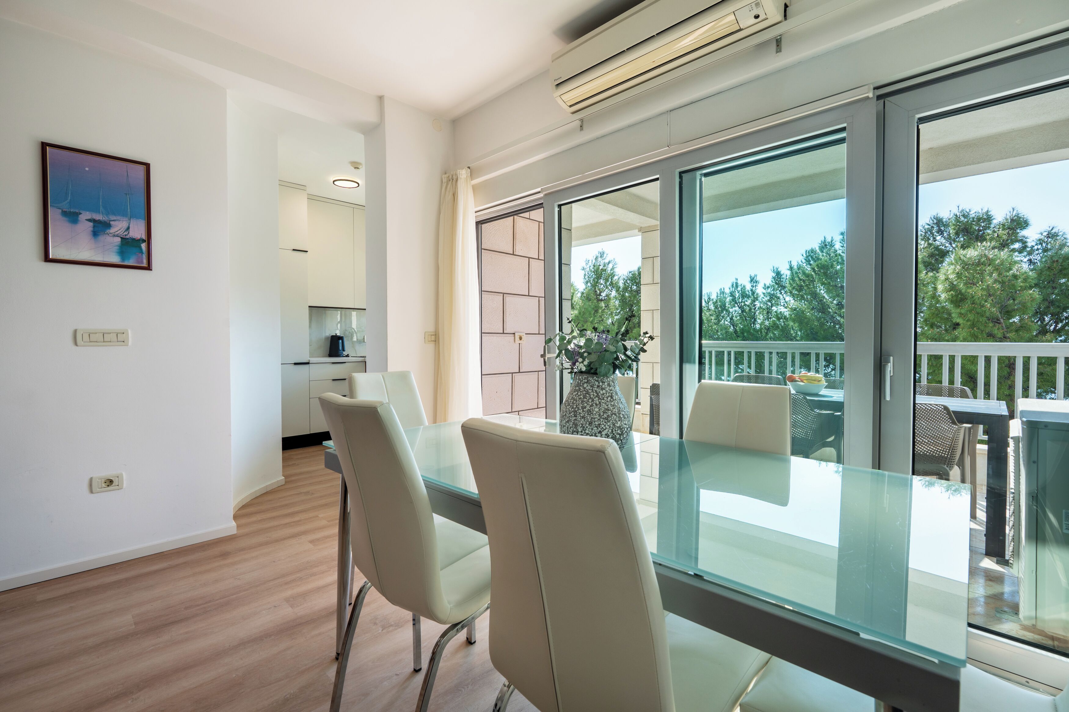 A modern living room with a glass dining table, comfortable chairs, a large window with a view of greenery, and a kitchen in the background.