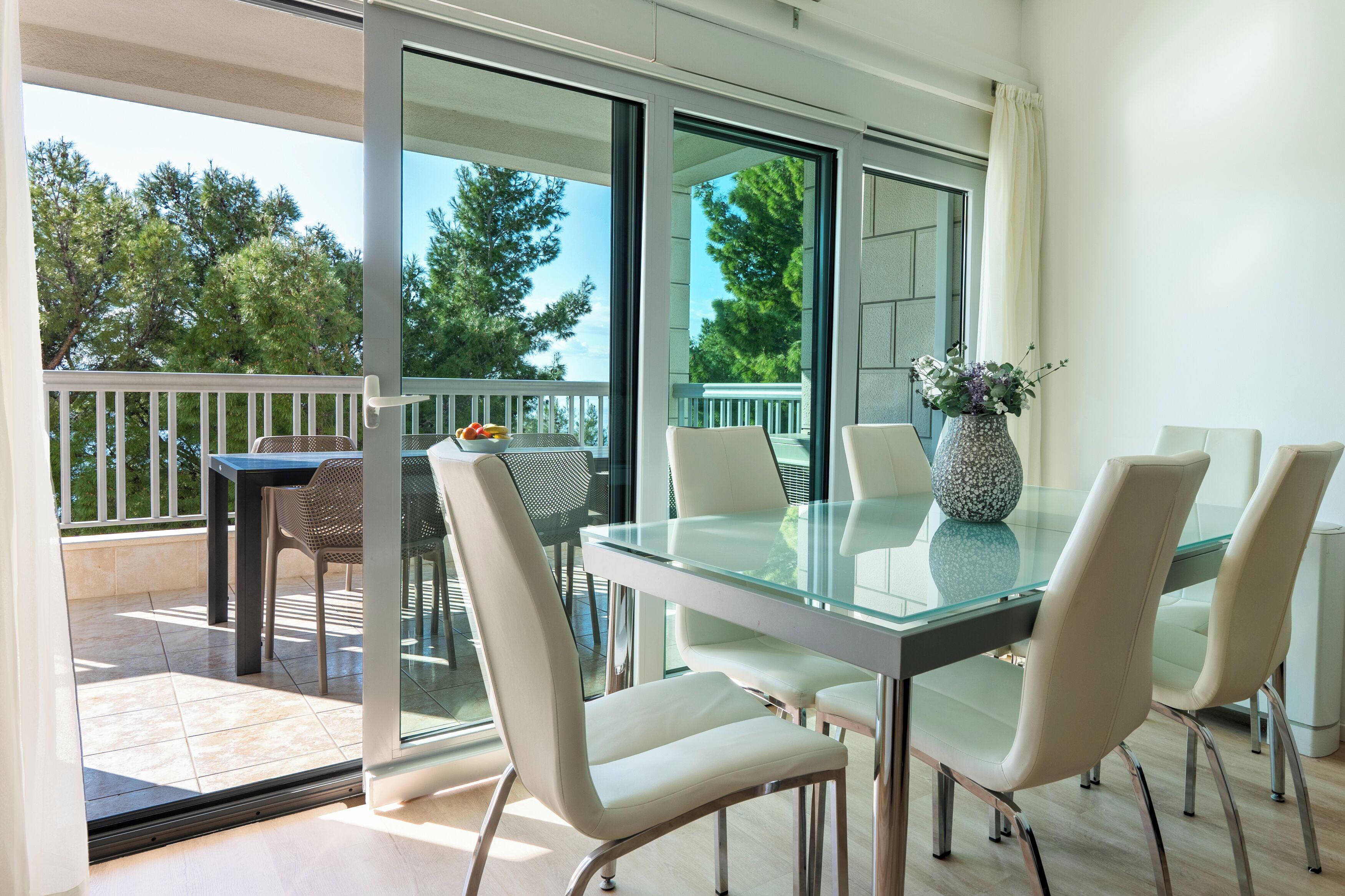 A modern dining area with a glass table and white chairs, large windows, and a view of trees.