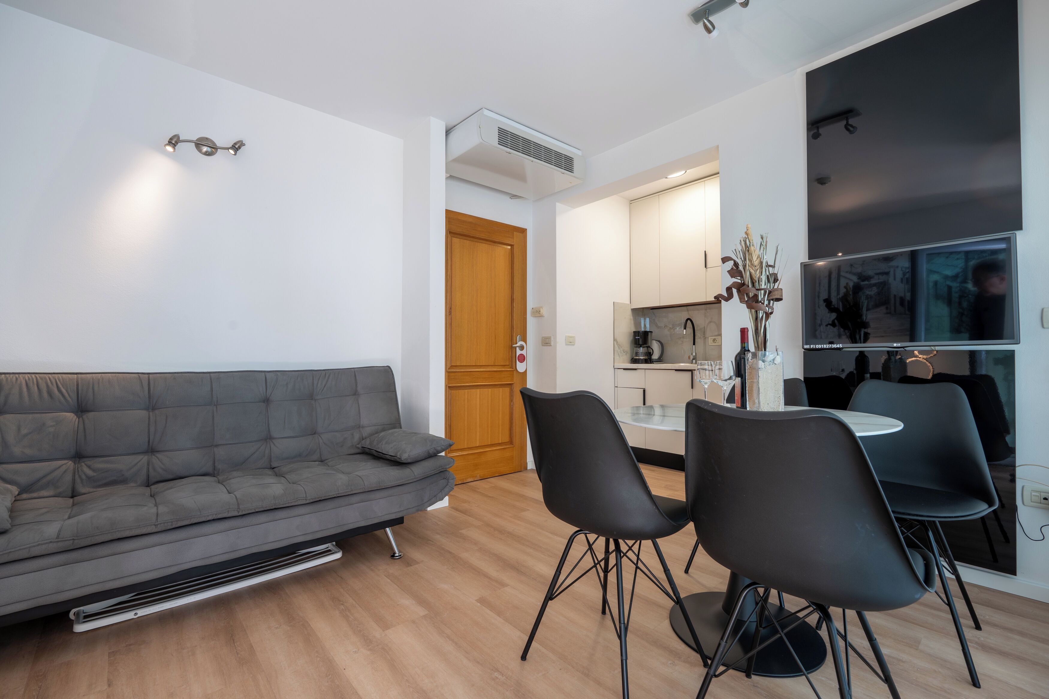 Un salon moderne avec un canapé gris, des chaises noires, une porte en bois et un coin cuisine équipé d’un téléviseur.