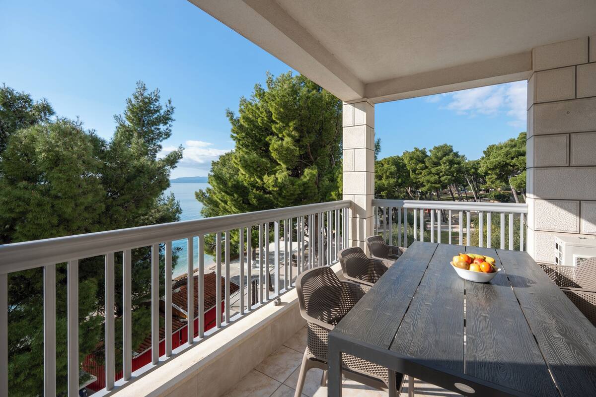 A balcony with a table and chairs, a bowl of fruit, and a view of the sea and trees.