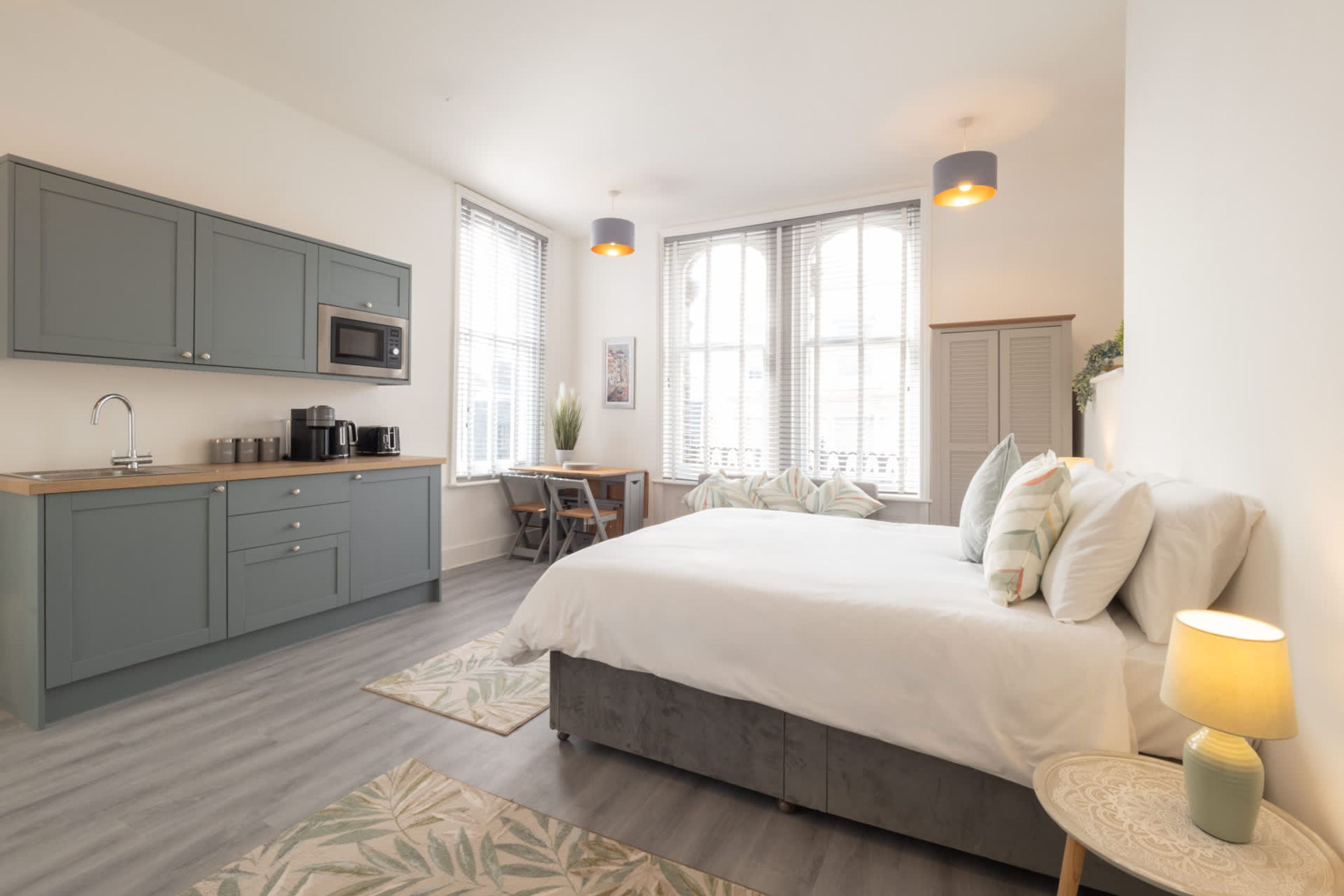 A modern bedroom with a large bed, a bedside table with a lamp, a desk with a chair, and a kitchenette with cabinets and a microwave.