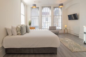 Une chambre avec un grand lit, deux lampes de chevet, un téléviseur fixé au mur et une fenêtre avec des stores.