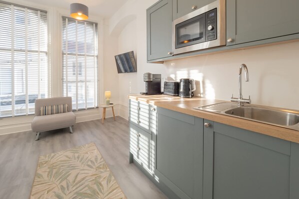 A modern kitchen with a built-in microwave, a coffee maker, and a sink. There is a small table with a lamp and a sofa in the adjacent living area. - Host Stay Harbour Rest (Ramsgate)