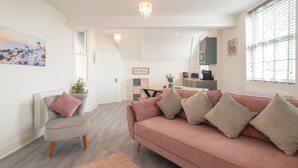 A modern living room with a pink sofa, a grey armchair, and a coffee table. There is a kitchen area in the background with a dining table and chairs. - Host Stay The Crow s Nest (Ramsgate)