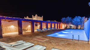 Área de piscina com espreguiçadeiras e piscina à noite.