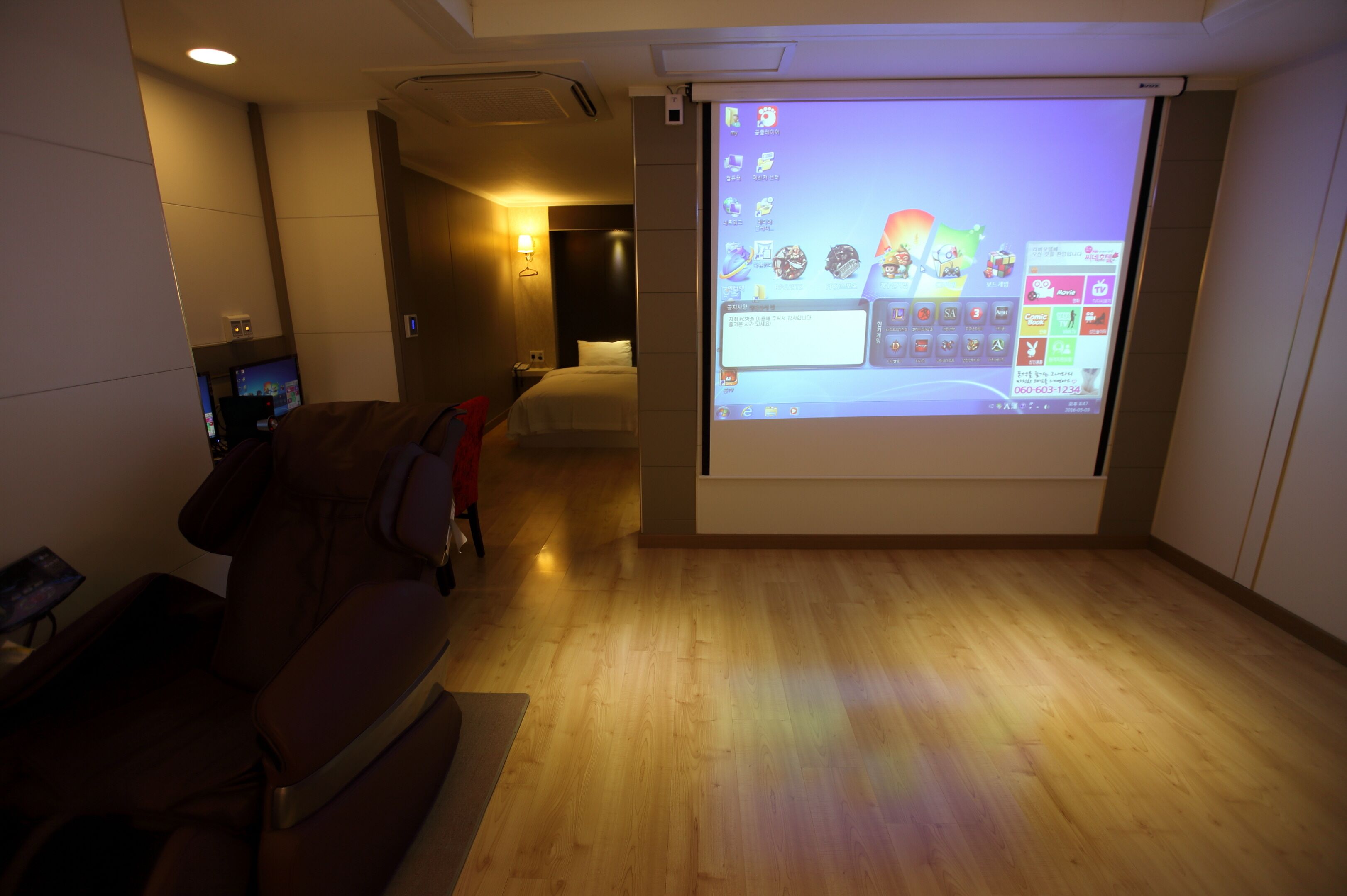 A hotel room with a large screen displaying a digital interface, a leather sofa, a wooden floor, and a bed with a headboard.