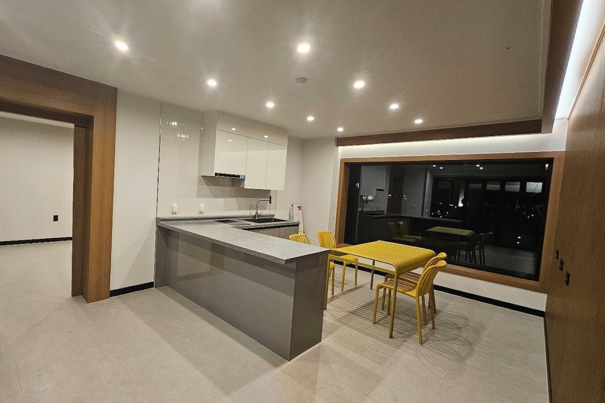 A modern kitchen with a central island, a dining area with yellow chairs, and a large window offering a view of the city at night.