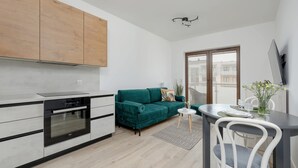 A modern kitchen with wooden cabinets, a built-in oven, a green sofa, a dining table with chairs, and a large window.