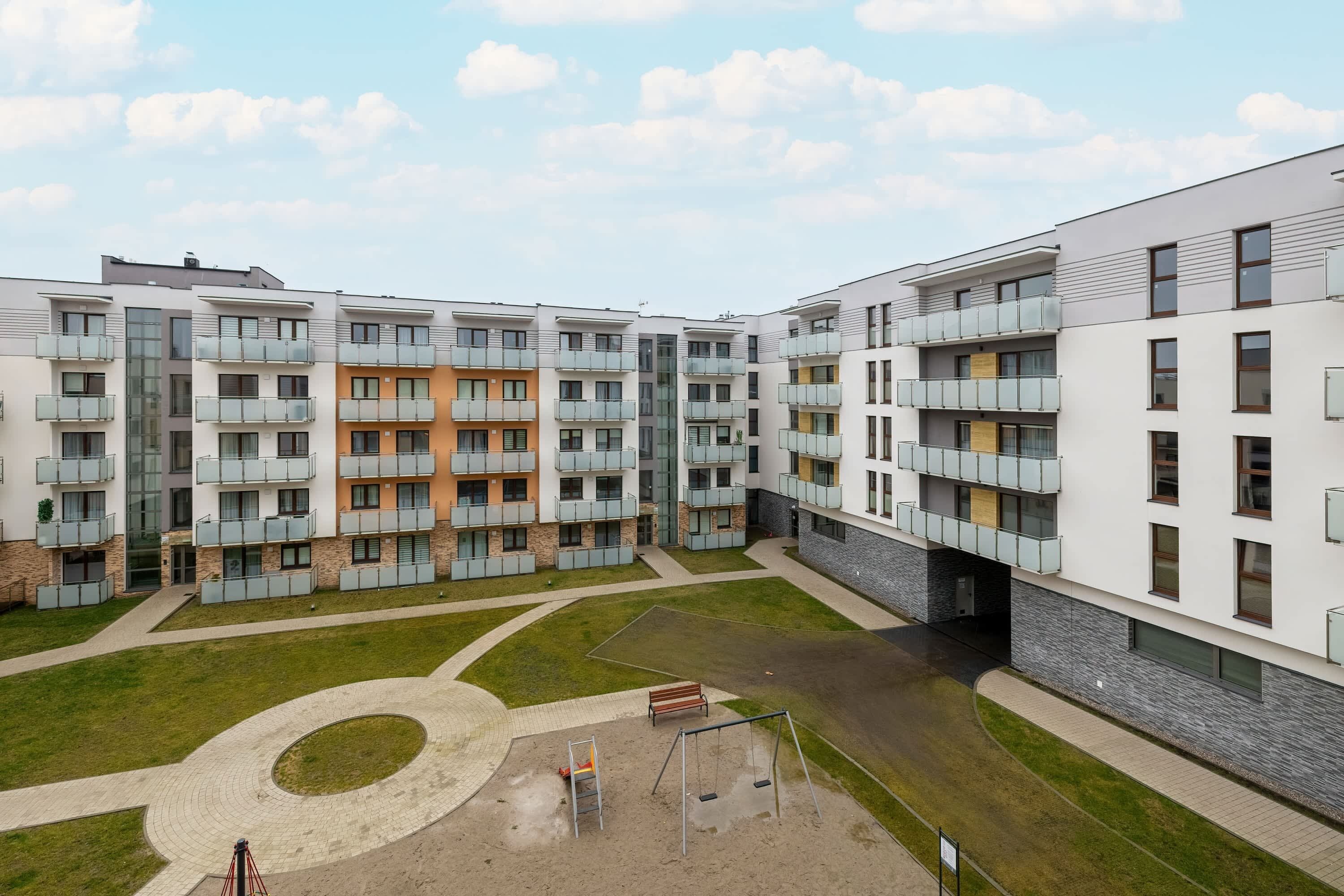 A modern apartment complex with multiple balconies, a central courtyard with a swing set, and a paved walkway.