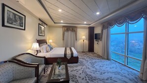 A hotel room with a large bed, two bedside tables with lamps, a sofa, a glass coffee table with a plant, and a large window offering a city view. - Hotel Royale de CASA (Guwahati)