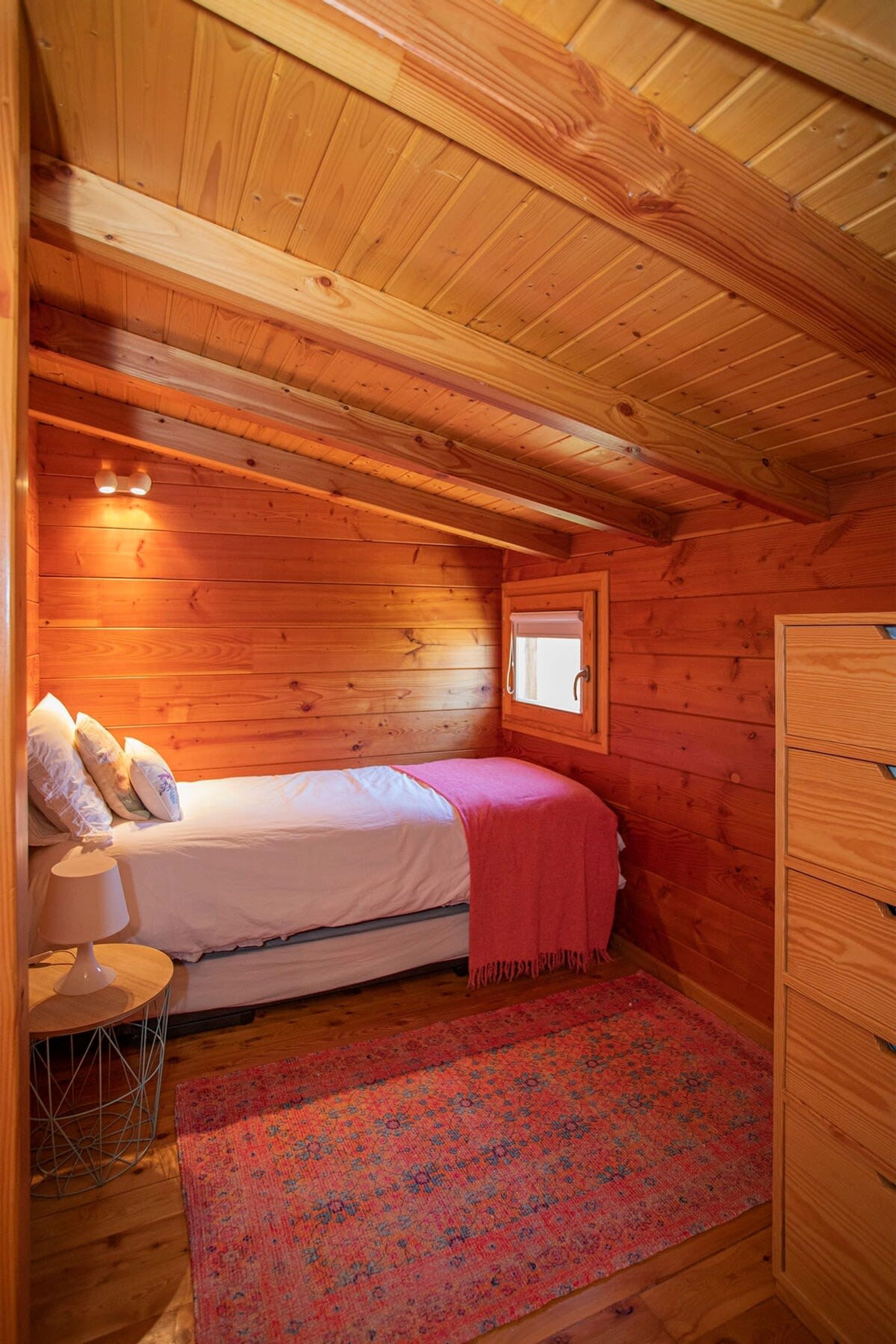 Un dormitorio de cabaña acogedor con techo de madera, una cama individual con almohadas, una mesita de noche con lámpara, una ventana con persiana y un aparador de madera.