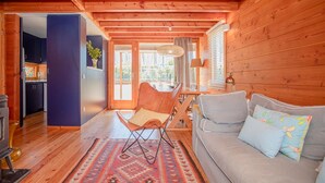 A cozy cabin interior with a wooden ceiling, a dining area with a table and chairs, a sofa with cushions, and a kitchen area visible through an open door.