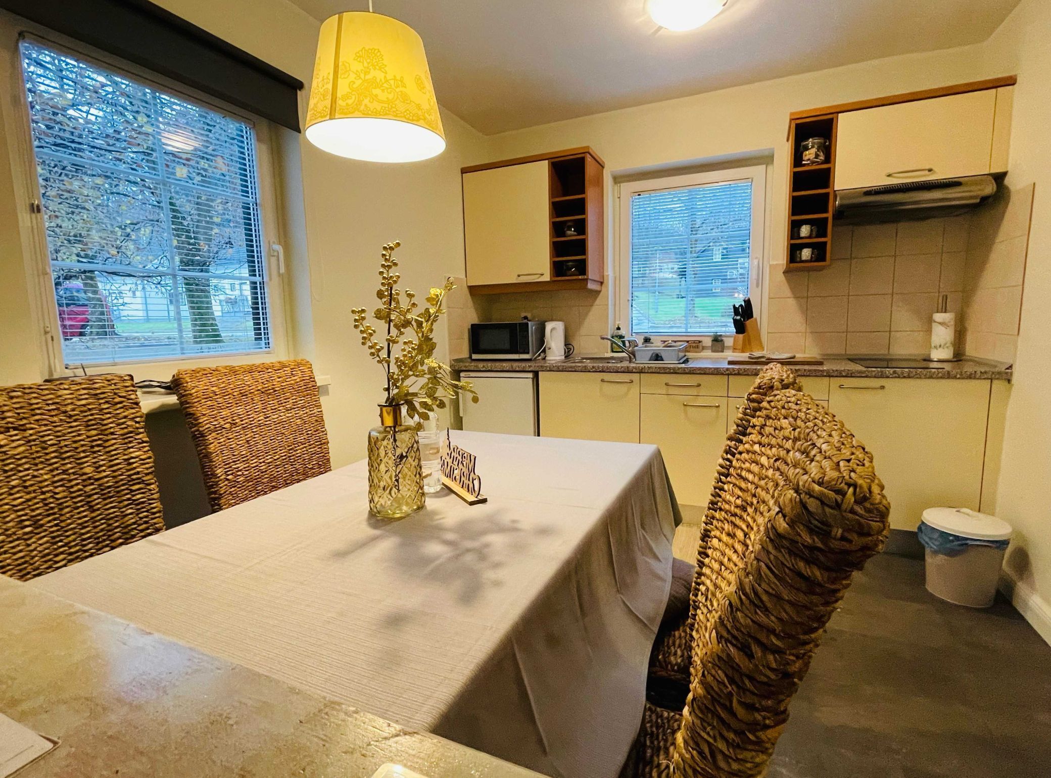 A compact kitchen with a dining area, featuring a table with a tablecloth, wicker chairs, and a hanging light fixture.