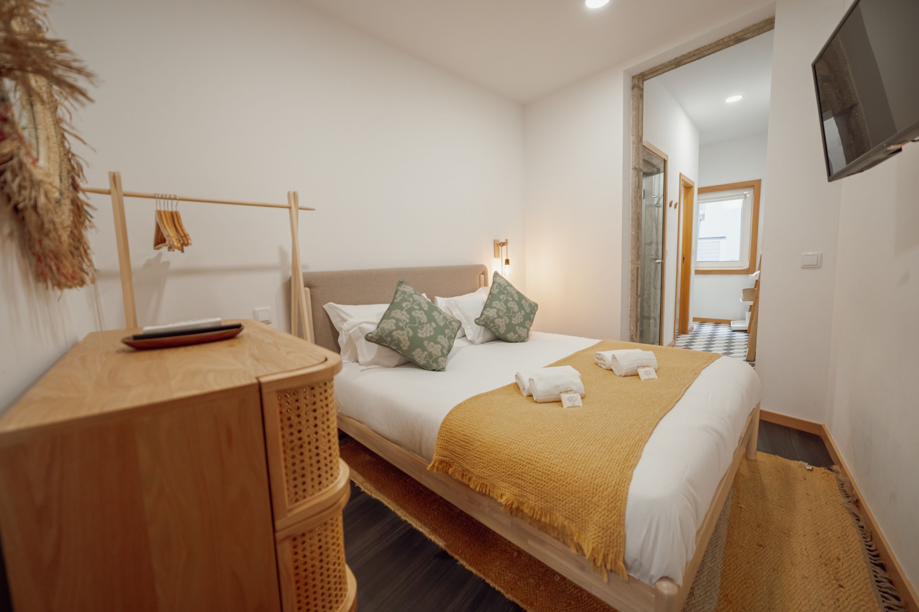 A hotel room with a bed, a wooden desk, a TV mounted on the wall, and a window with blinds.