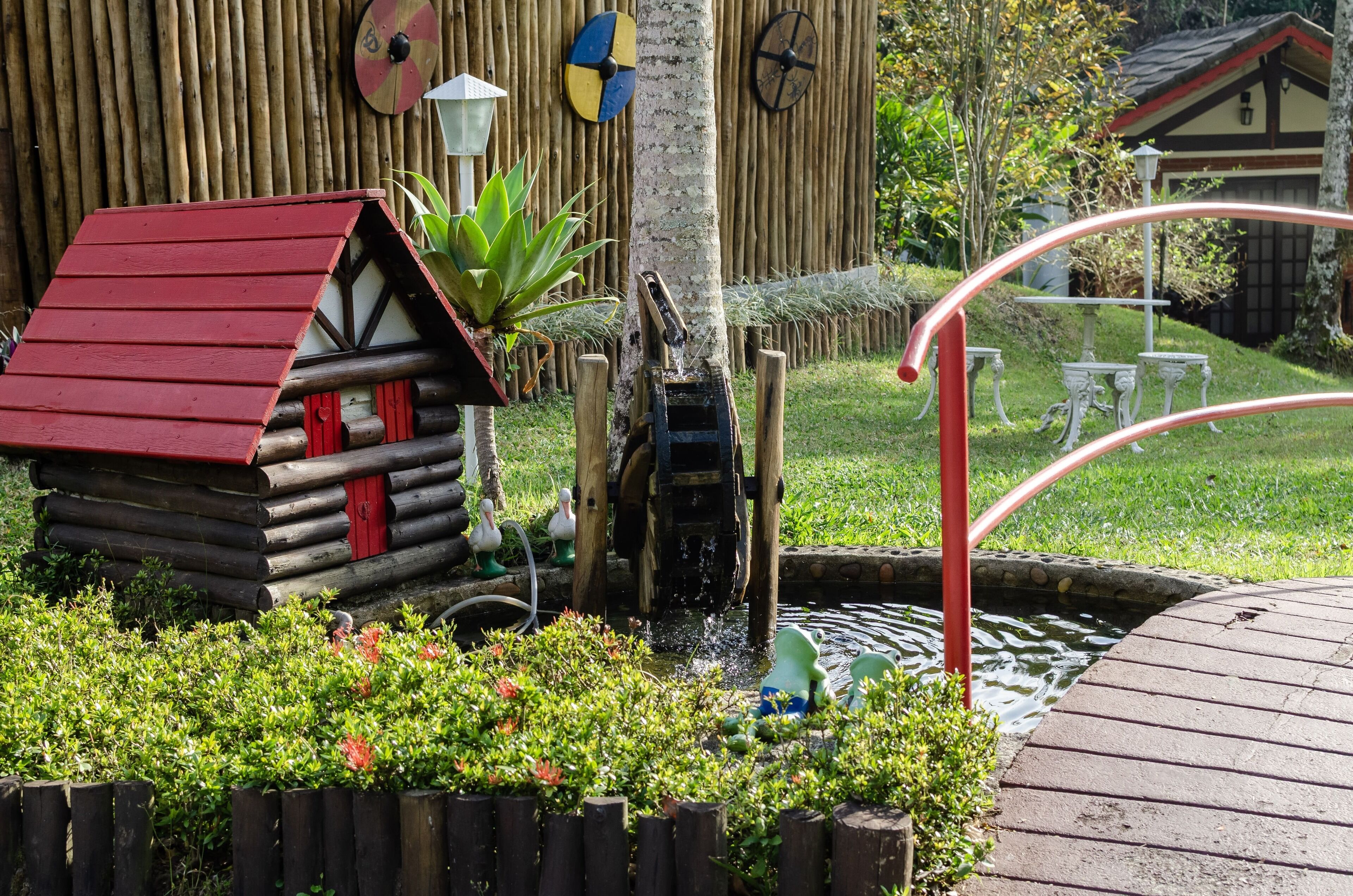 Una cabaña de madera con techo rojo, un estanque pequeño con fuente de agua y una cerca de madera.
