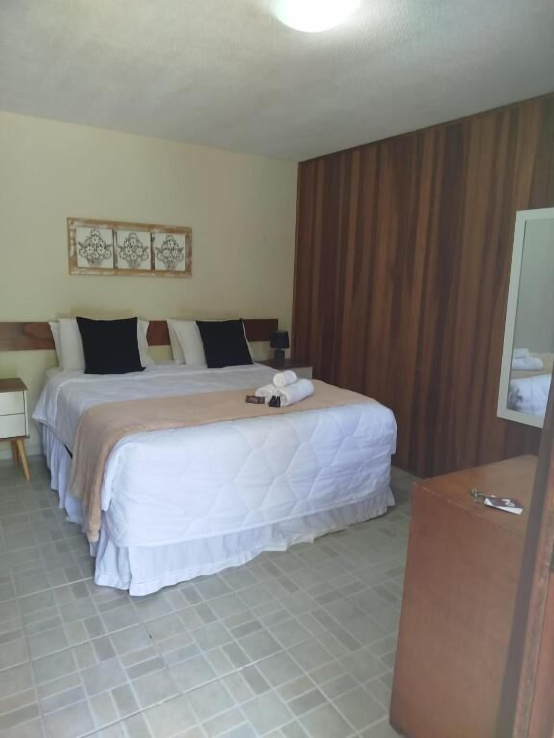 Quarto de hotel com cama grande, duas mesas de cabeceira, cabeceira de madeira, pequena escrivaninha e um espelho.