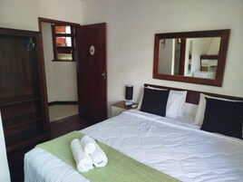 Una habitación de hotel con una cama, una manta verde, dos toallas enrolladas, un armario de madera, una mesita de noche con una lámpara y un espejo en la pared.