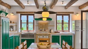 Una cocina con armarios verdes, una mesa de comedor de madera con un bol de fruta y vistas al exterior a través de ventanas amplias.