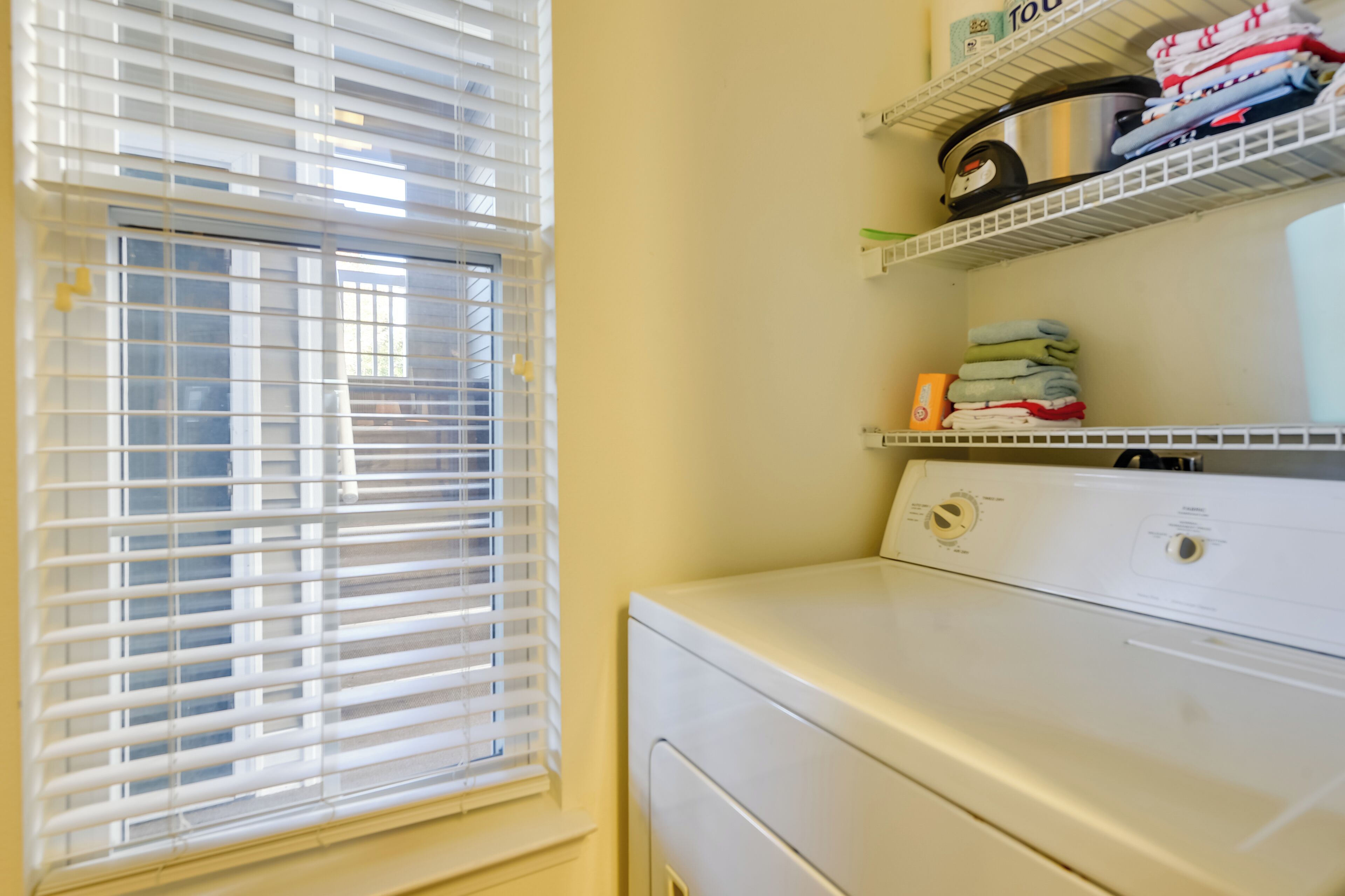 A laundry area with a front-loading washer and dryer, a window with blinds, and shelves with folded clothes and laundry supplies.
