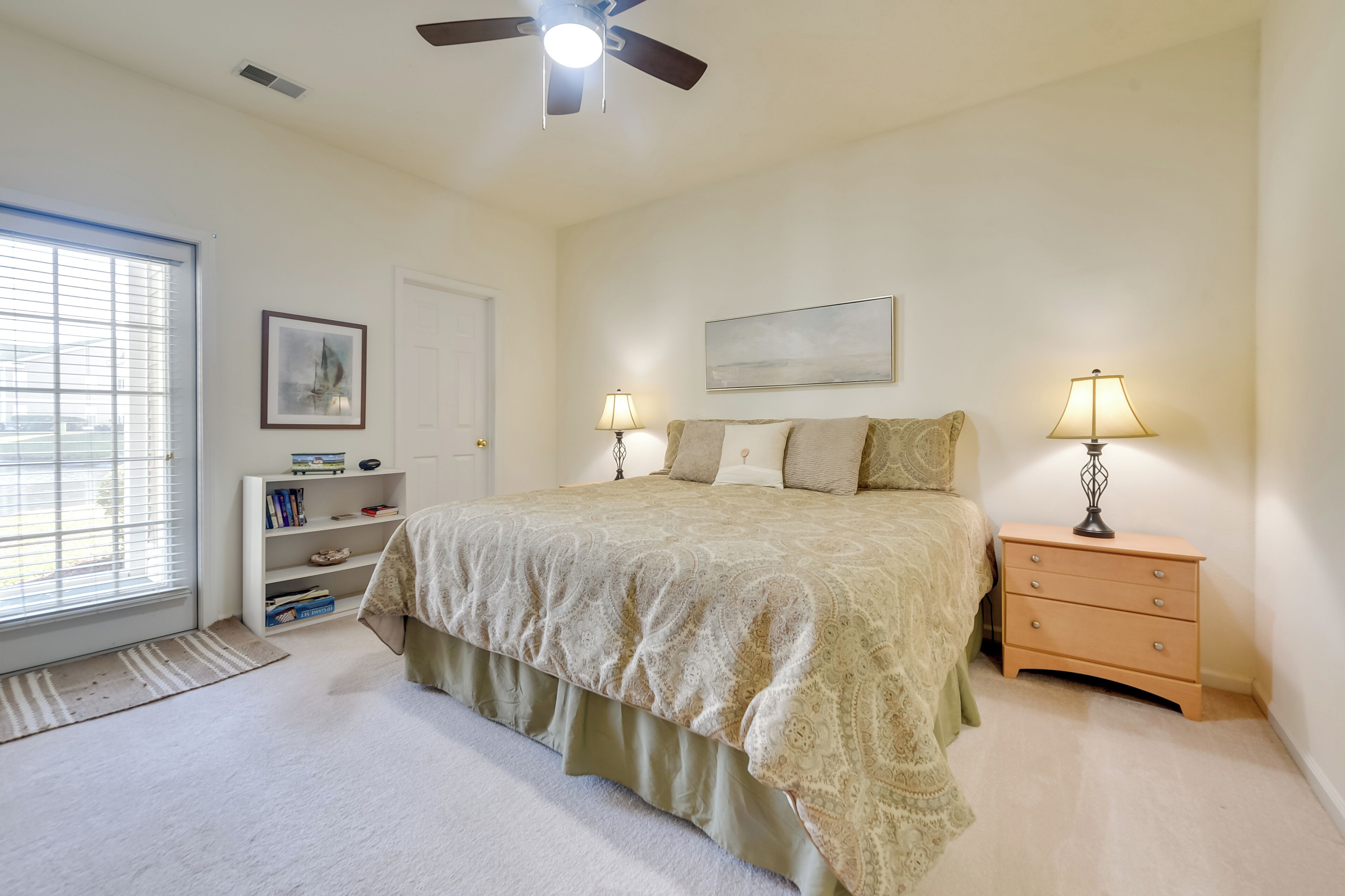 A bedroom with a bed, two bedside tables with lamps, a bookshelf, a door, and a window with blinds.