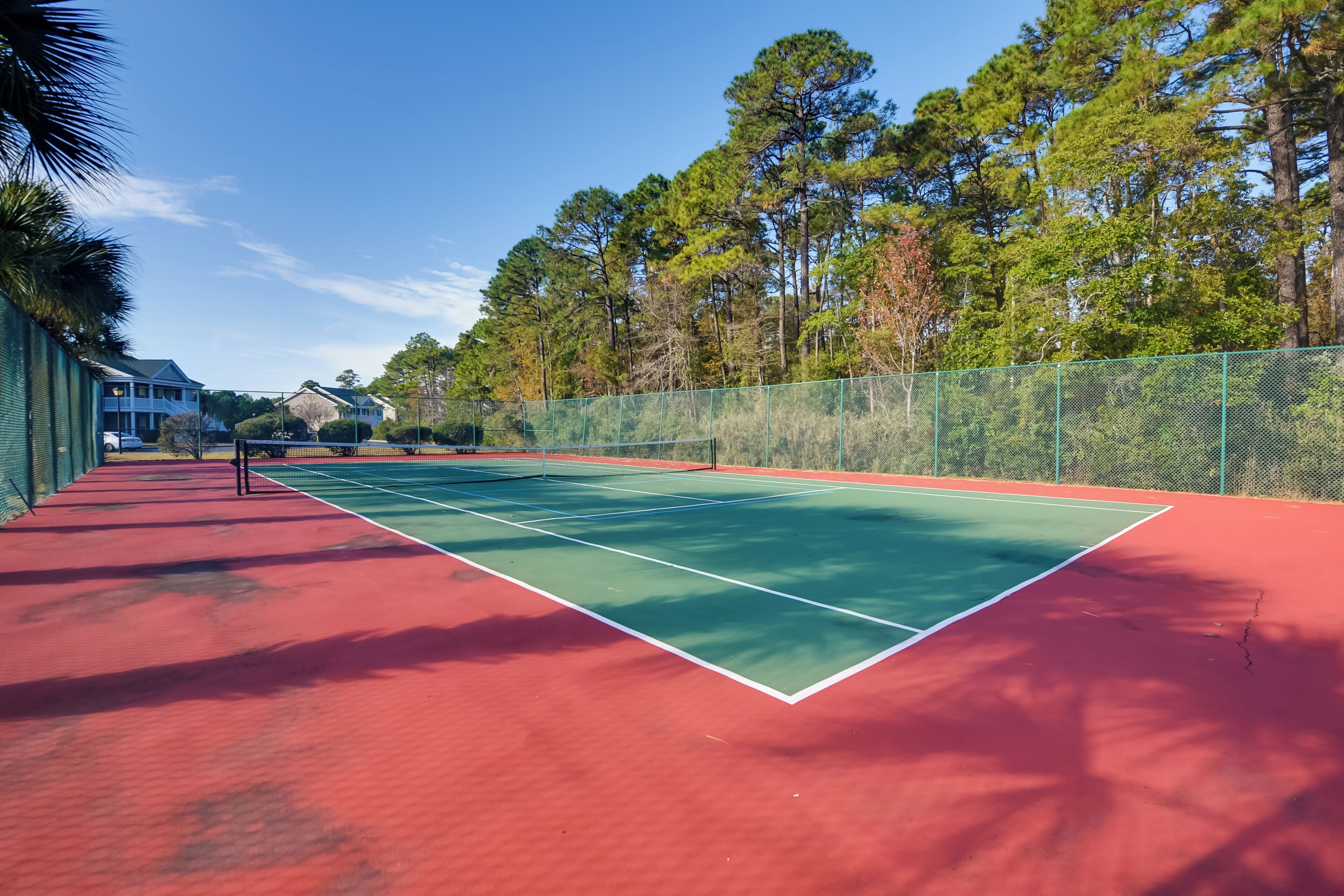 Een tennisbaan met een rode ondergrond, omringd door een groene hekken en bomen.