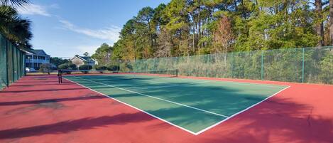 Een tennisbaan met een rode ondergrond, omringd door een groene hekken en bomen.