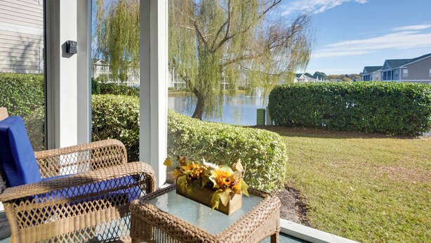 A wicker chair and table set on a balcony with a view of a lake and a weeping willow tree. - Pond Views & Community Perks: Sunset Beach Condo! (Sunset Beach)