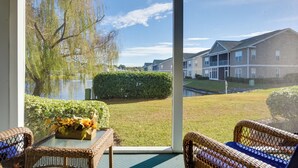 A balcony with wicker furniture, a view of a lake, and residential buildings in the background. - Pond Views & Community Perks: Sunset Beach Condo! (Sunset Beach)