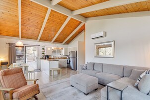A spacious living area with a high wooden ceiling, a grey sofa, a wooden chair, and a kitchen in the background.