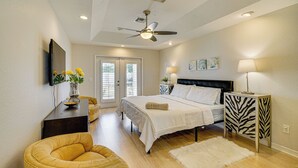 Une chambre à coucher comprenant un lit, un ventilateur de plafond, une table de chevet avec une lampe, une chaise et une porte donnant sur une autre pièce.