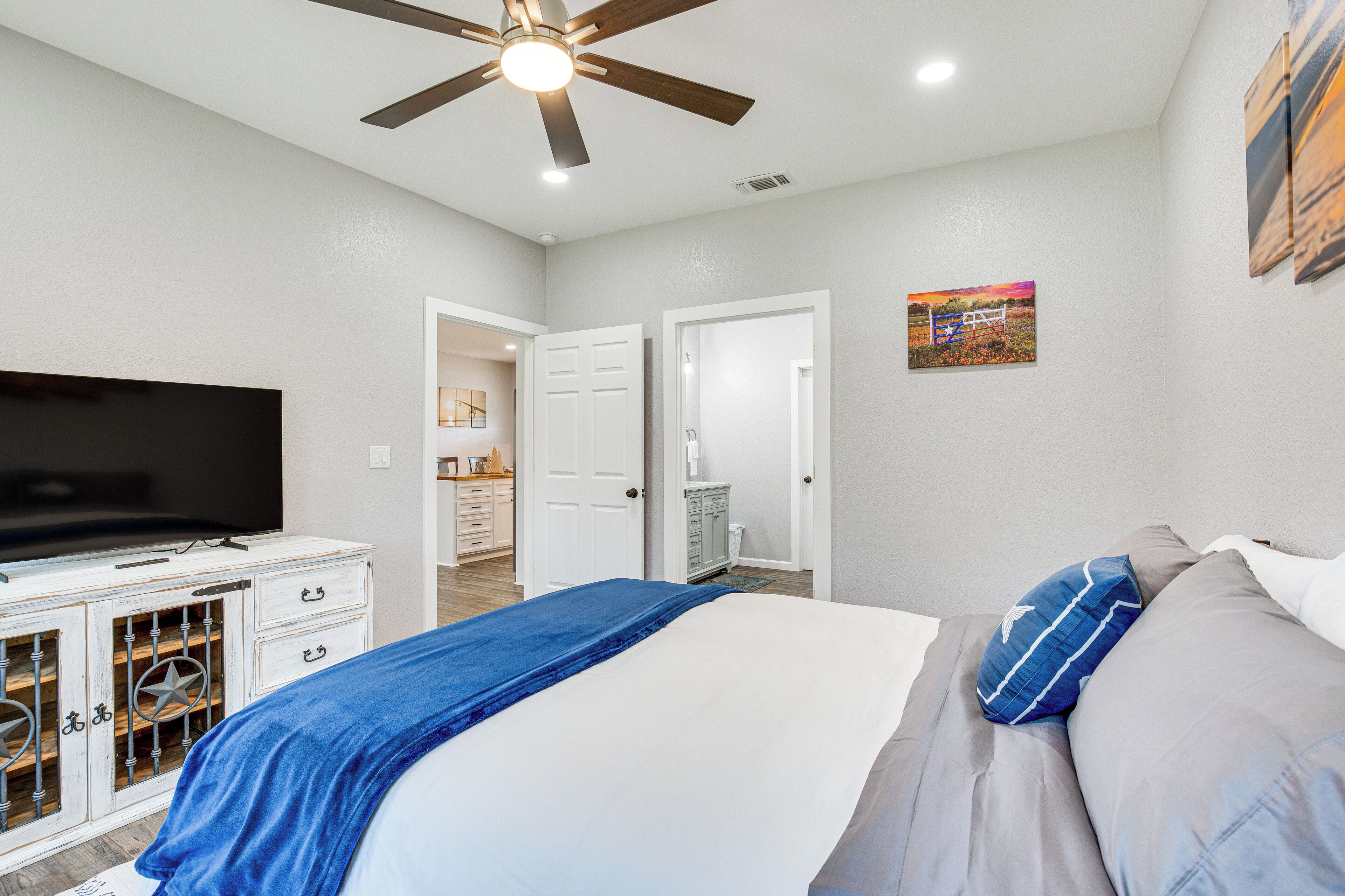 A bedroom with a bed, a TV, a ceiling fan, and a door leading to a bathroom.