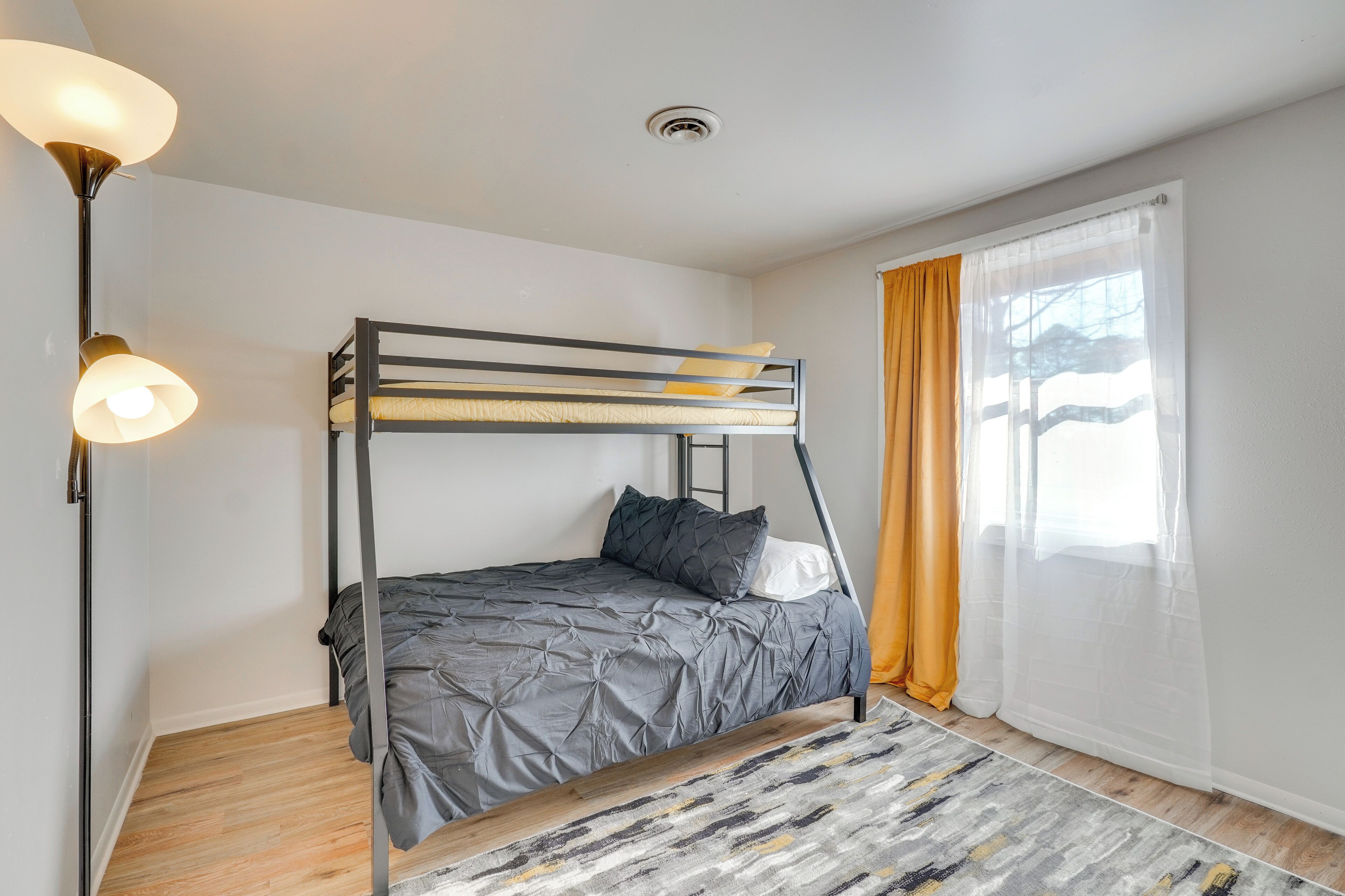 Een slaapkamer met stapelbed, hoofdbord, vloerlamp, een rug met patroon en een raam met gordijnen.