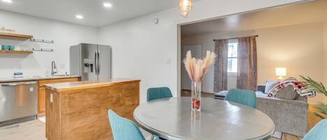 Una cocina moderna con una isla central, un comedor con sillas color esmeralda y una sala de estar con sofá y lámpara.
