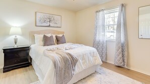 Ein Schlafzimmer mit Bett, Nachttisch, Lampe, Deckenventilator, Fenster mit Vorhängen und einem Wandbild.