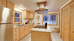 Una cucina con mobili in legno, un frigorifero in acciaio inossidabile, un piano di lavoro in granito, un fornello a gas, una finestra vista alberi e un ventilatore a soffitto.