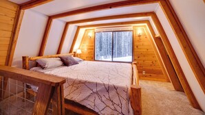 Una camera da letto mansardata accogliente, con travi a vista in legno, un’ampia finestra vista su una foresta innevata, un letto con copripiumino decorato e un piccolo comodino con una lampada.