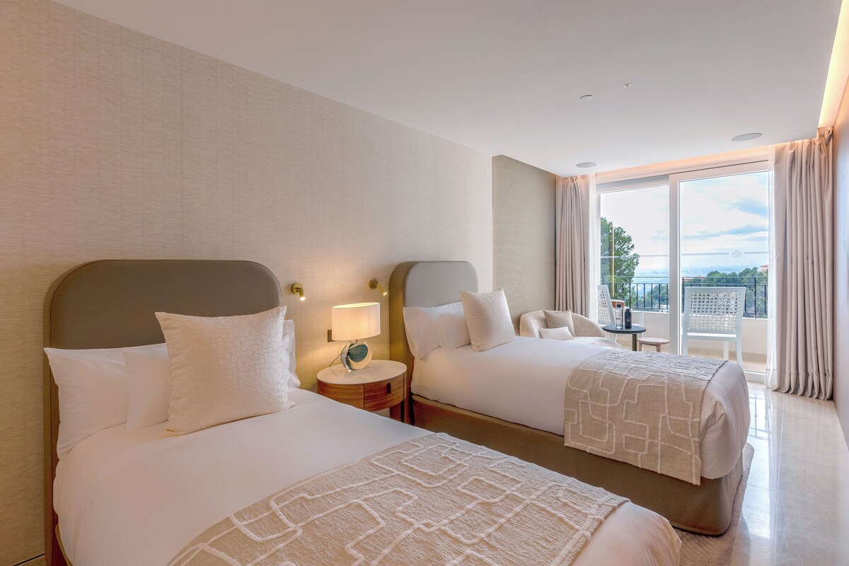 A hotel room with two beds, a round table, a chair, and a balcony with a view.