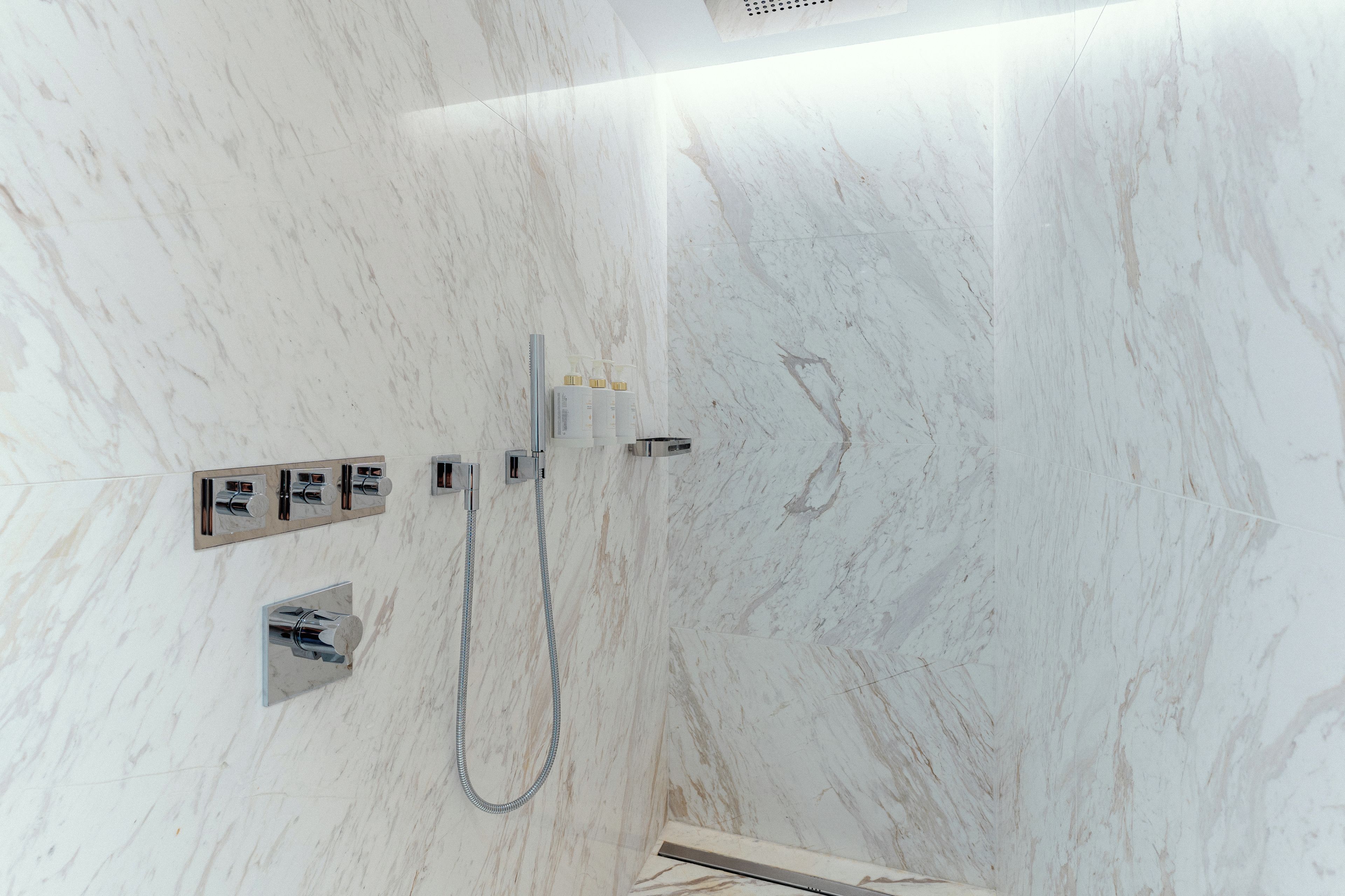 A bathroom with a marble wall, a shower with a handheld showerhead, and a control panel with various buttons and a water temperature control.