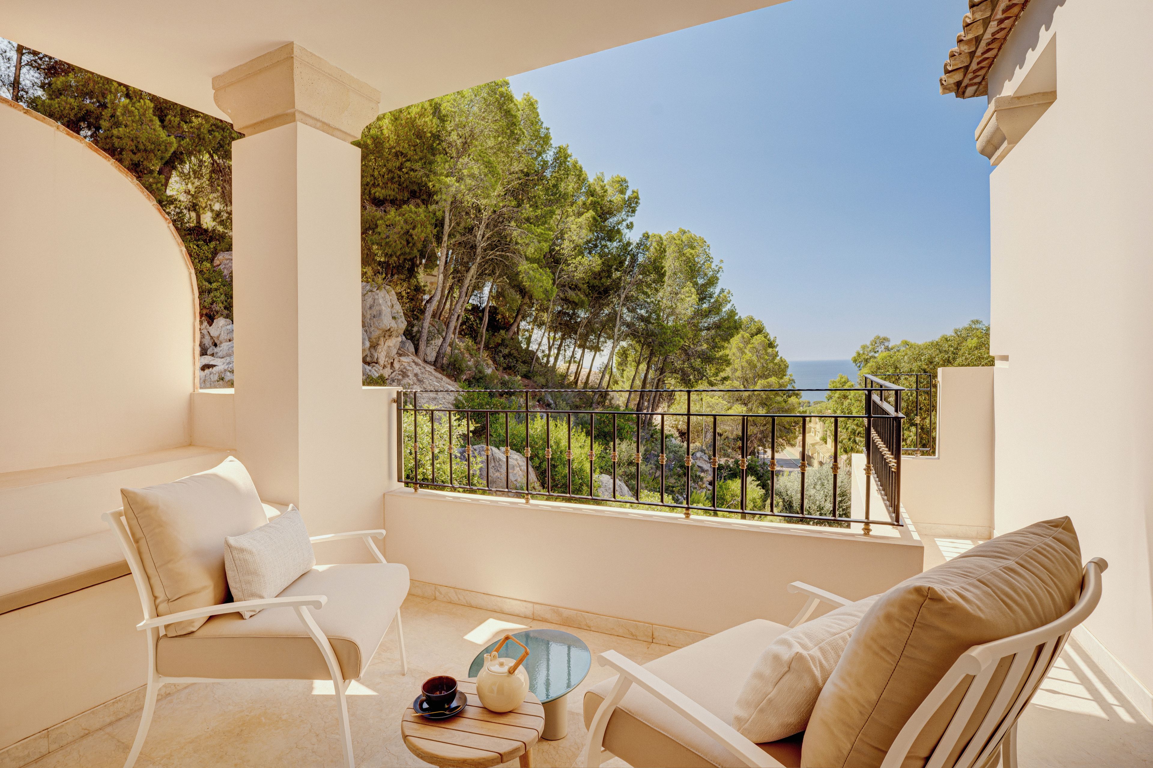 Ein Balkon mit weißer Möbel, ein kleiner Tisch mit Teekanne und Tasse, und Blick auf Bäume und Meer.