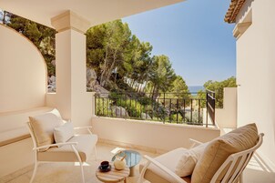 A balcony with white furniture, a small table with a teapot and cup, and a view of trees and the sea.