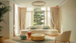 Un salon moderne avec un canapé incurvé, une table basse ronde et un fauteuil. On y trouve une grande fenêtre avec des rideaux, un lustre et une plante en pot.