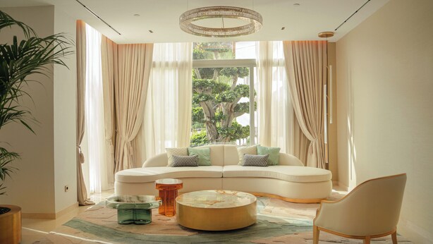 A modern living room with a curved sofa, a round coffee table, and a chair. There is a large window with curtains, a chandelier, and a potted plant. - ZEM Wellness Clinic Altea - 5 Stars Grand Luxury (Altea)