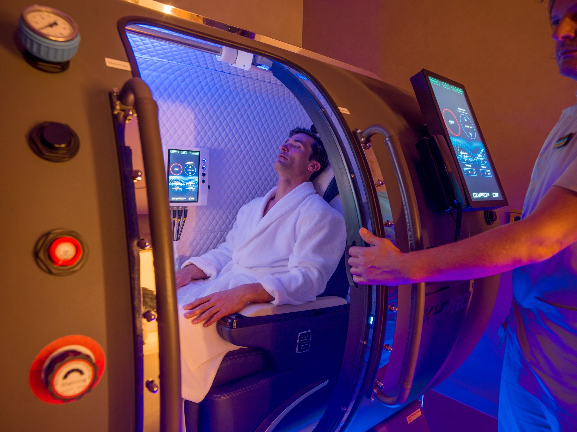 A person in a white robe inside a treatment room with a digital display and control panel.