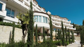 Un hôtel à plusieurs étages doté d’un balcon incurvé, de murs en pierre et d’un jardin aménagé avec des cyprès.