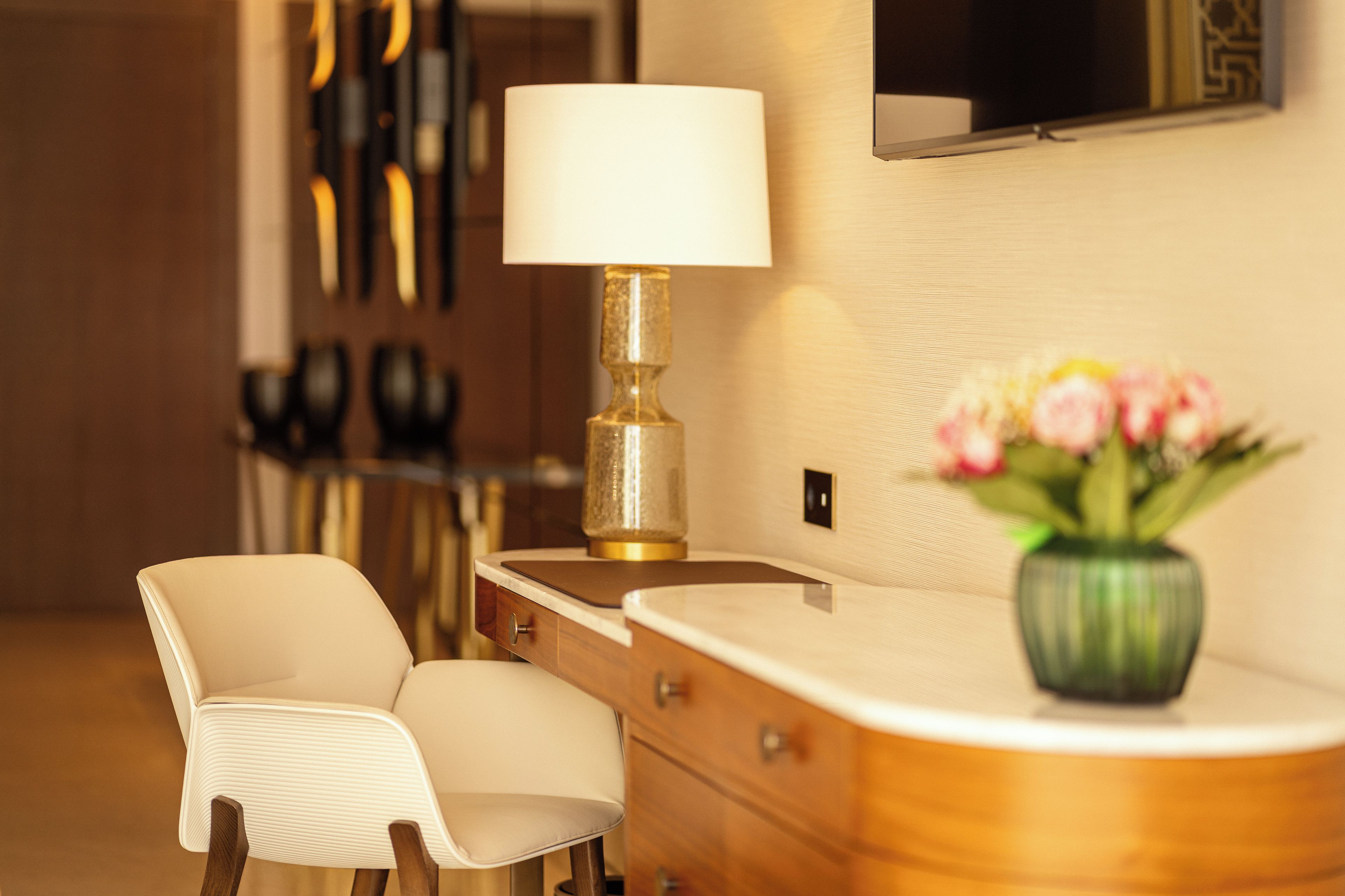 A hotel room with a desk, a chair, a lamp, a vase with flowers, and a mirror on the wall.