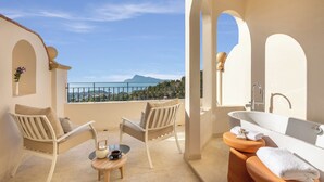 Un balcon avec deux chaises blanches, une petite table ronde et une vue sur une montagne et la mer.