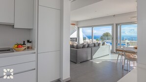 A modern kitchen with white cabinets, a dining area with a grey sofa, and a view of the sea through large windows. - Villa YANSA by Abahana Villas (Benitachell)